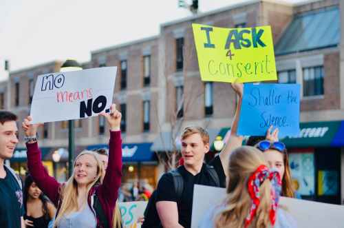 Student protestors march down Spruce St. protesting sexual violence. One white female students holds up a sign saying "no means no" on a white background. Another white male student holds up a yellow sign with the words "I ask 4 consent." Another student holds up a blue sign that reads "Shatter the Silence."