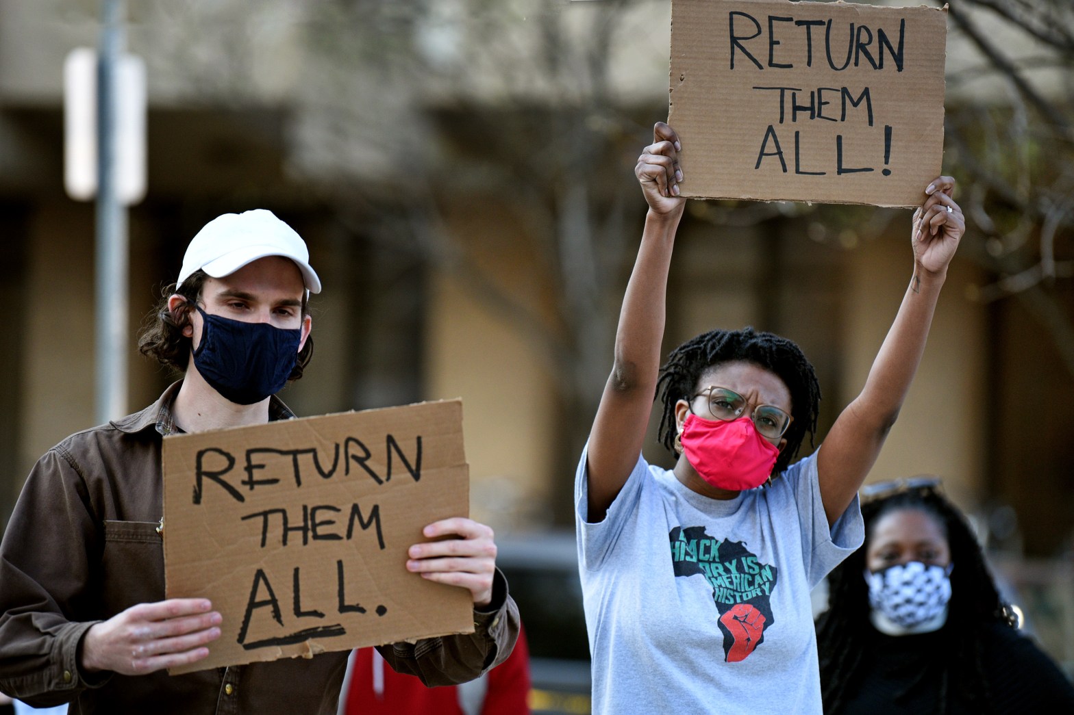 Two community members protest Penn's possession of the Morton Collection of skeletons from enslaved people. A white man with dark hair and a Black woman hold signs that read "RETURN THEM ALL."