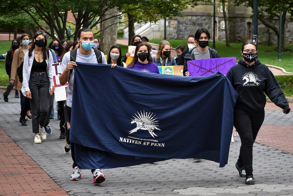 Students from Natives at Penn march down Locust Walk. Two students hold a navy colored banner with the Natives at Penn logo on it.