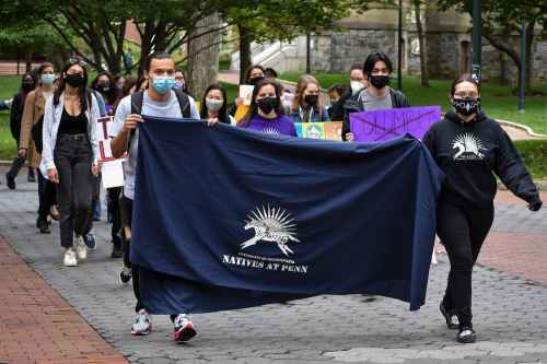 Students from Natives at Penn march down Locust Walk. Two students hold a navy colored banner with the Natives at Penn logo on it.