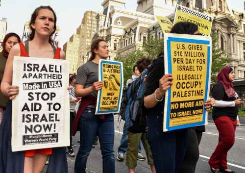 Philadelphians march down a street during a rally calling for an end to Israel's occupation of Palestine. On the left side of the photo, a white person in a red dress is carrying a sign that reads "Israeli apartheid, made in U.S.A. Stop aid to Israel now! Mundo Oberero Workers World Party." In the middle of the photo, another white person in a grey shirt is carrying a sign with a yellow background and black texts that reads "Resistance is justified when people are occupied." On the right side of the photo, a third protestor is carrying a sign covering their face, also with a yellow background and black text that reads "U.S. gives $10 million per day for Israel to illegally occupy Palestine. No more aid to Israel now!" Other protestors, a couple wearing red hijabs, are also marching in the background.