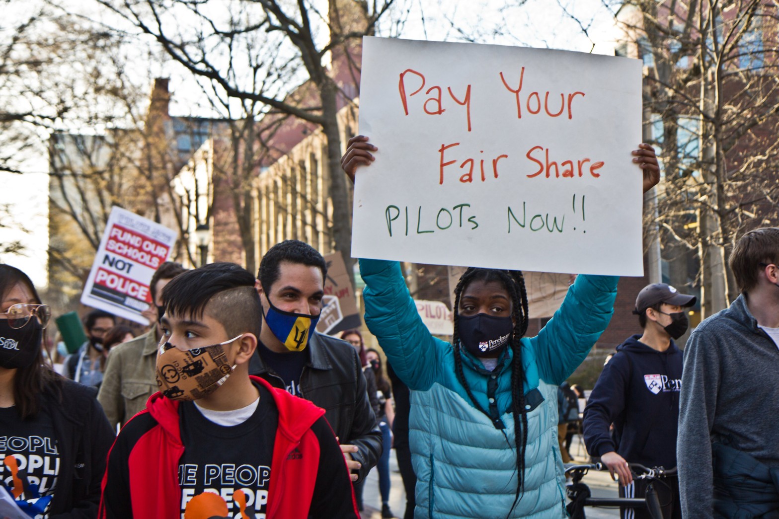 Protesters march on the campus of the University of Pennsylvania demanding the school and Drexel University pay PILOTS to support Philadelphia schools on March 30, 2021. A black student in a blue coat holds up a sign that says, "Pay your fair share PILOTs now!!" Another protestor in the background holds a sign that reads, "Fund our schools not police."