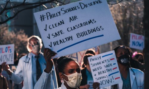 Protestors hold up signs advocating for PILOTs. A masked black woman with a black bun holds up a sign that says, "#PhillySchoolsDeserve ✔️ Clean Air, ✔️ Safe Classrooms, ✔️ Healthy Buildings." Another Black woman in a white coat and black hair down holds a sign that reads, "Philly students need safe + healthy schools now!" In the background are other protestors with signs that say, "PILOTs now!"